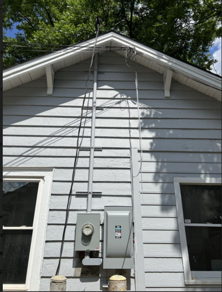 Electrical box and meter on a gray house exterior, with wires and a ladder-like structure ascending to the roof.