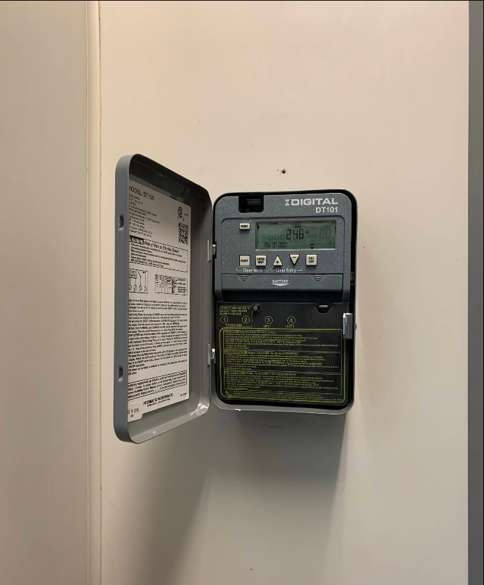 Grey irrigation timer on a wall, door open showing controls and instructions.