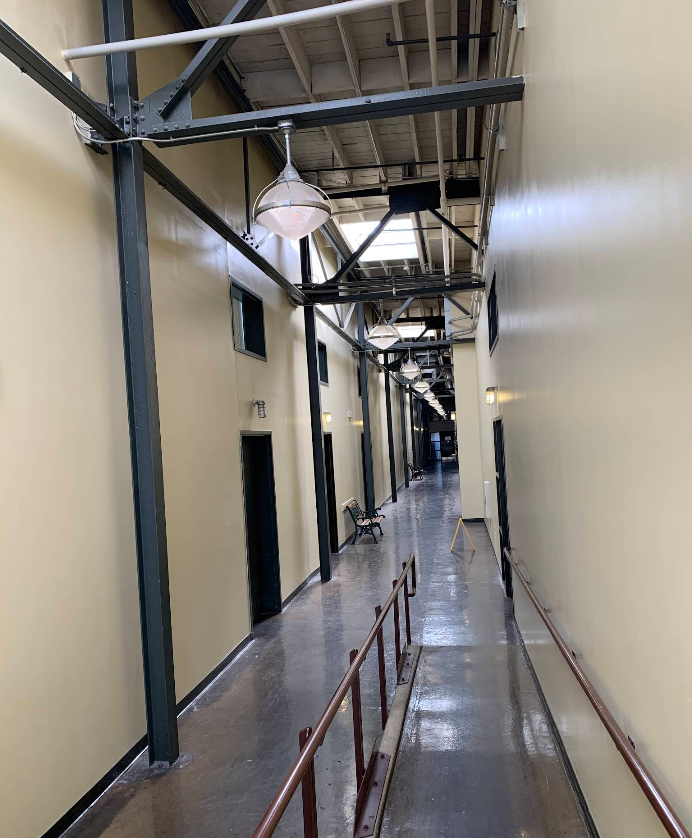 Long, narrow hallway with tan walls, black doors, and metal beams overhead.
