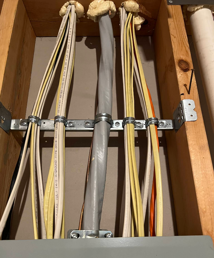 Electrical wires in a wooden frame, secured by metal brackets and clamps, above a gray panel.