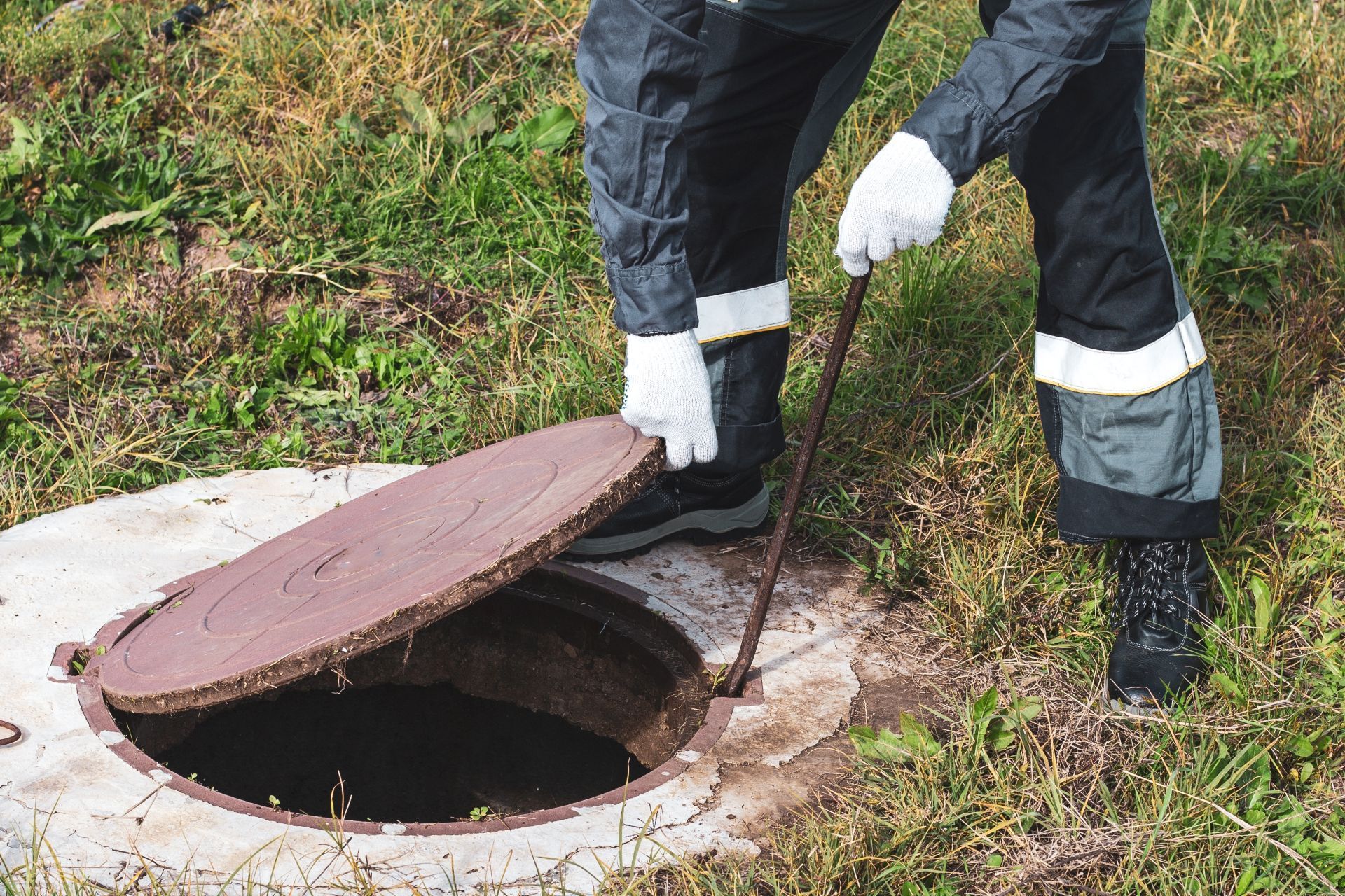 A person in uniform and white gloves uses a metal hook to lift a heavy circular cover off a ground-level manhole.
