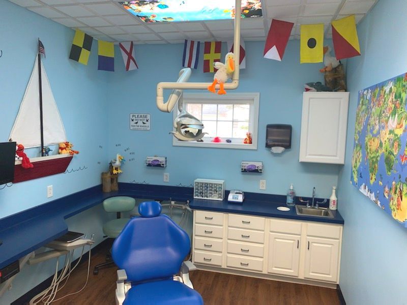 Colorful pediatric exam room with blue chair, white cabinets, nautical flags, map, and a small window.
