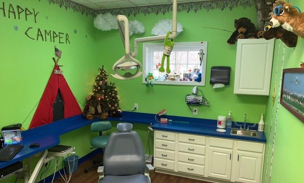 Bright green pediatric exam room with colorful chairs, cabinets, and a “Happy Camper” wall mural
