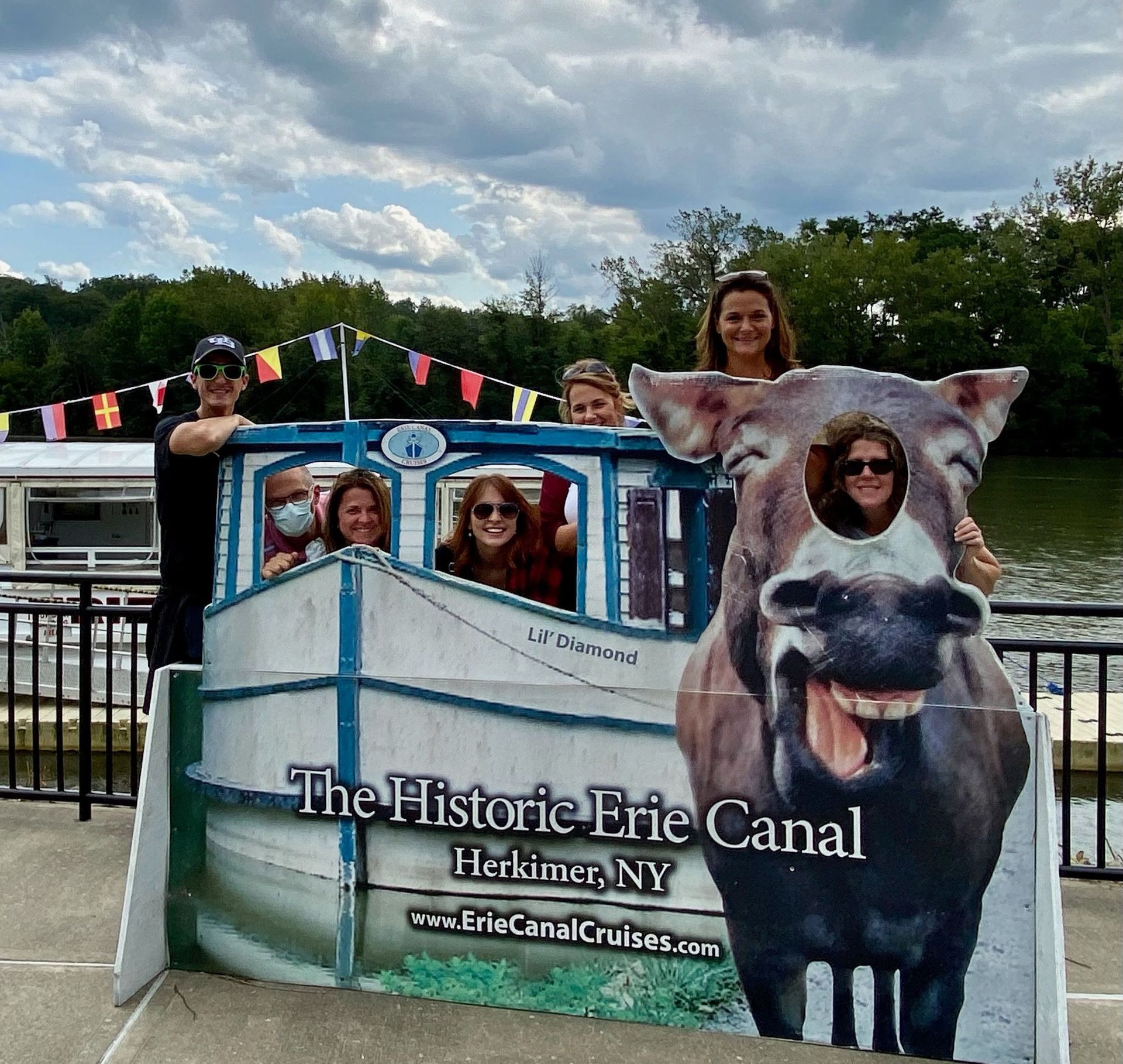 People posing behind a Historic Erie Canal cutout sign by the water on a cloudy day