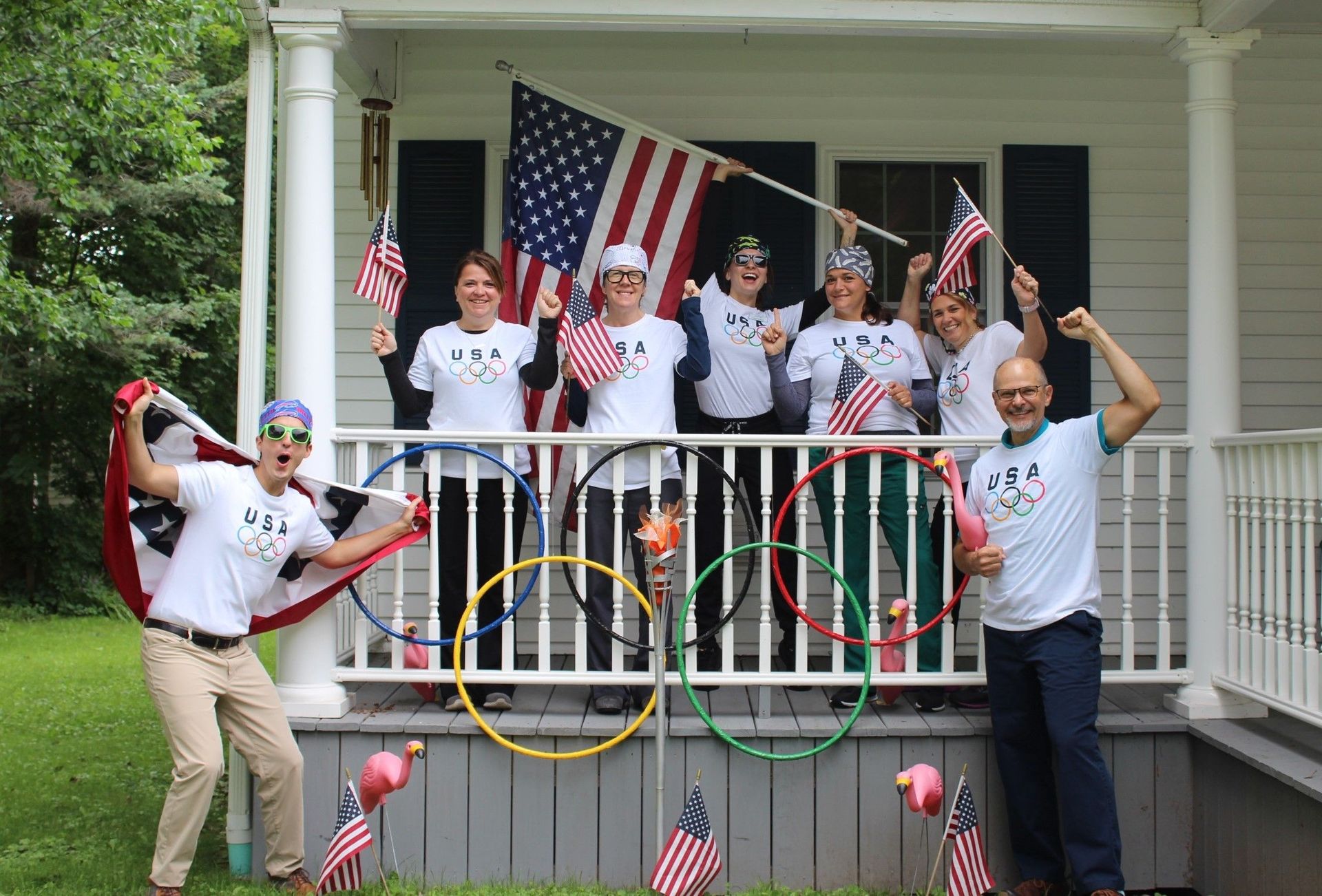 Group of people on a porch decorated with U.S. flags and Olympic rings, smiling and waving at the camera