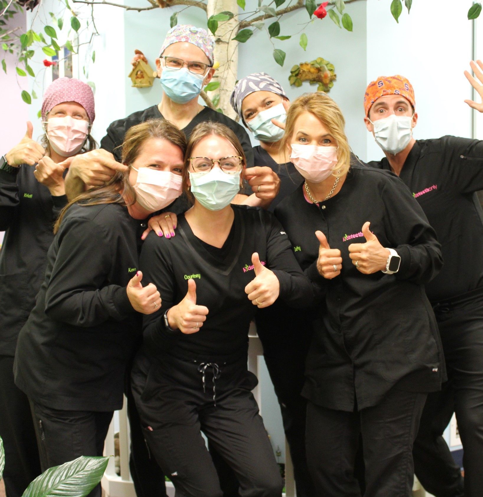 Six masked people in black scrubs posing indoors with thumbs up and smiles