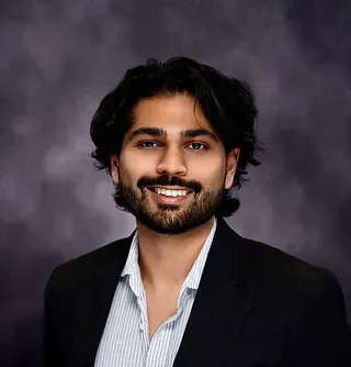 Portrait of a smiling person in a black blazer and light striped shirt against a gray backdrop