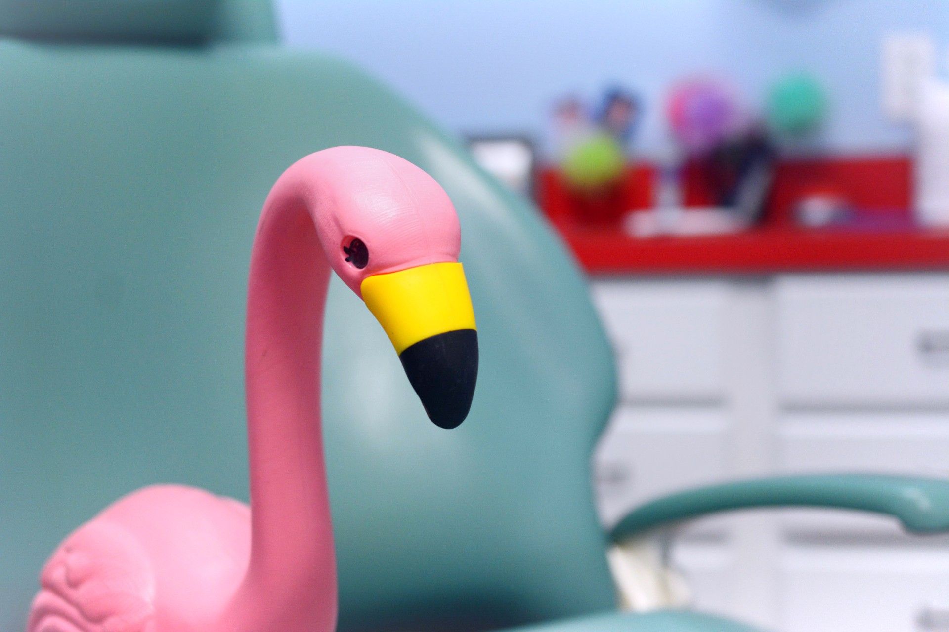 Pink flamingo toy with a yellow-and-black beak in front of a teal chair and blurred colorful toys.