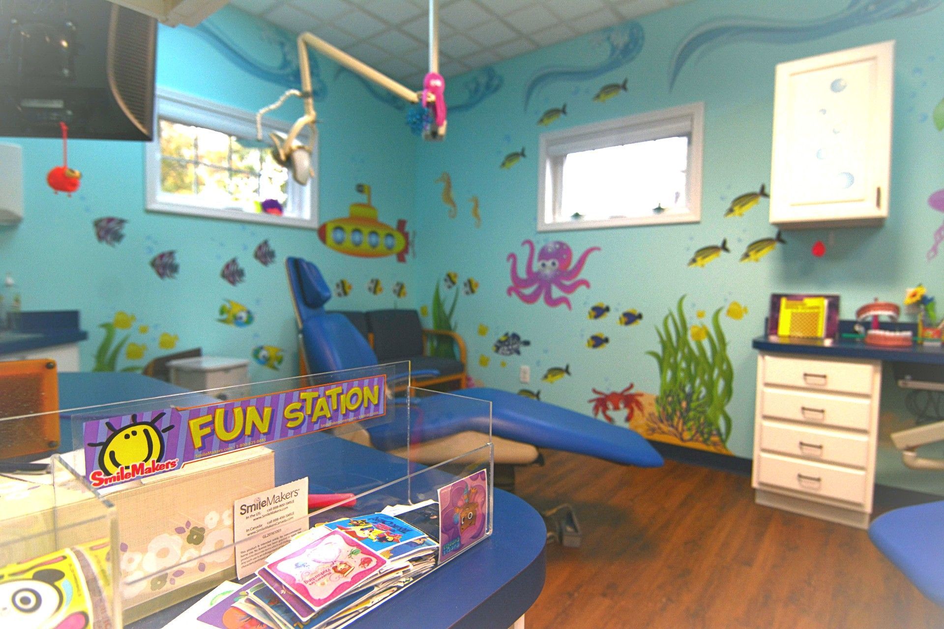 Colorful daycare playroom with a toy station, blue cushions, wall murals, and a cabinet near windows.