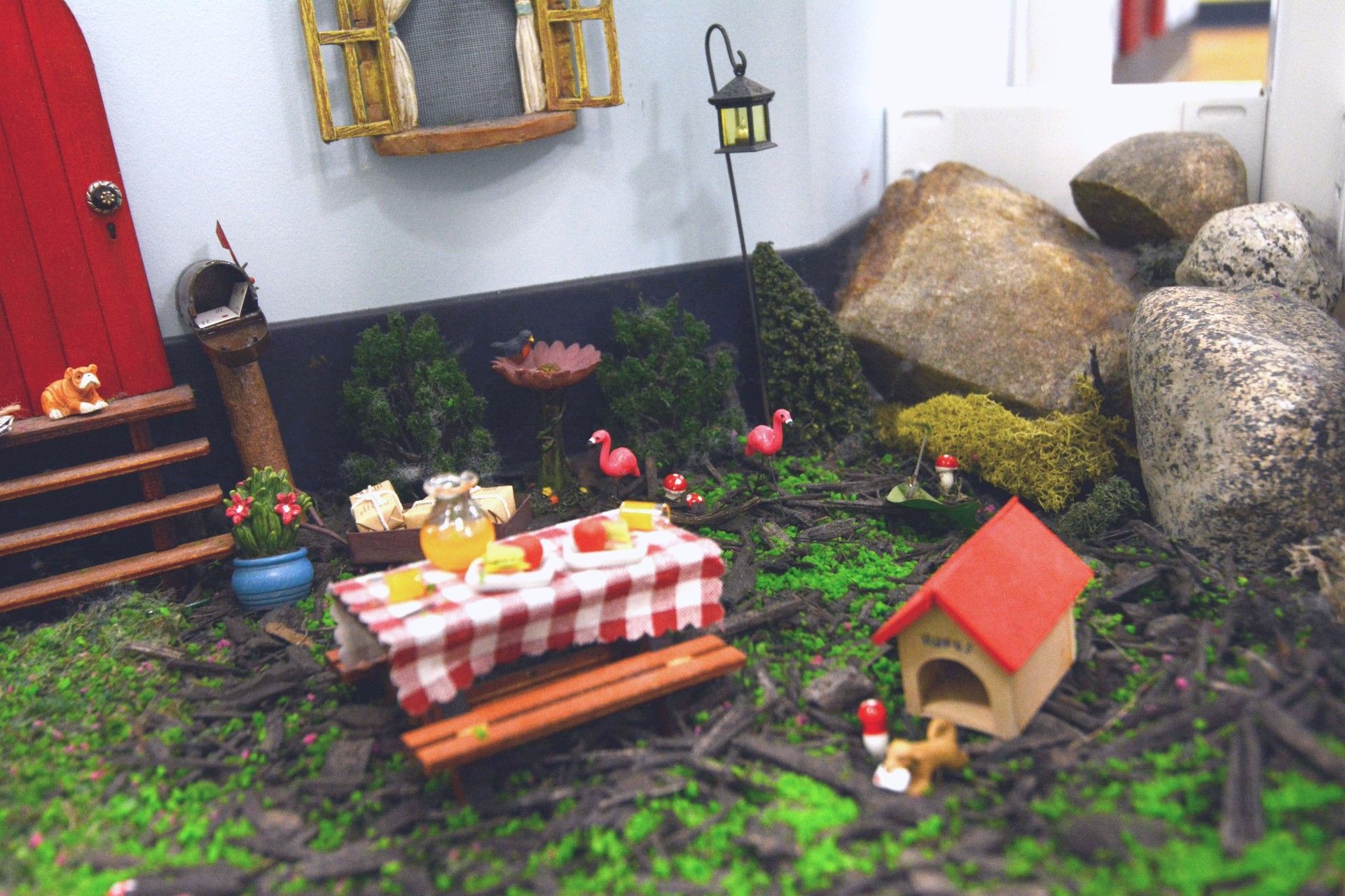 Miniature garden scene with a picnic table, red-roof birdhouse, flowers, and large rocks by a house wall