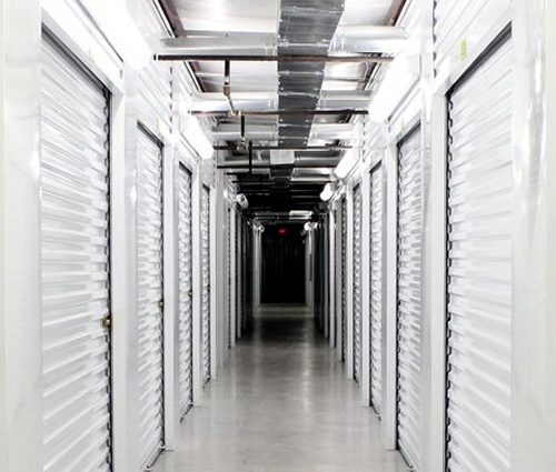 Hallway of storage units with white doors, metal ceiling fixtures, and a concrete floor.