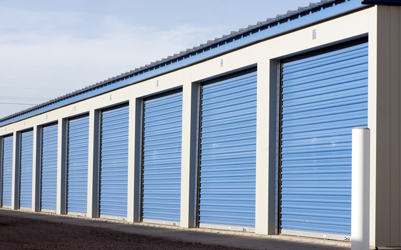 Row of blue storage unit doors under a blue roof, white frames, asphalt ground.