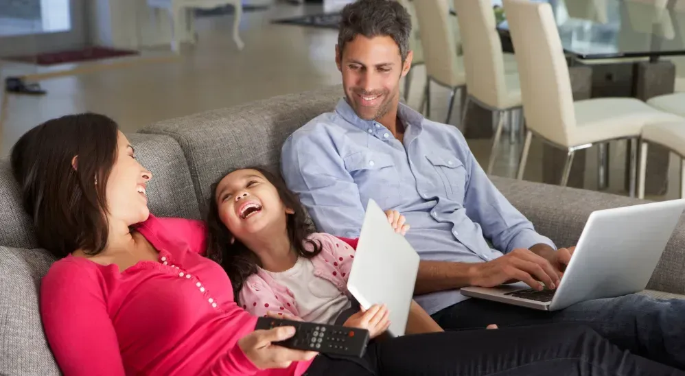 A family is sitting on a couch with a laptop and a remote control.