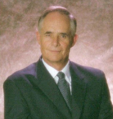 Man in a dark suit and tie, smiling slightly, against a muted, warm background.