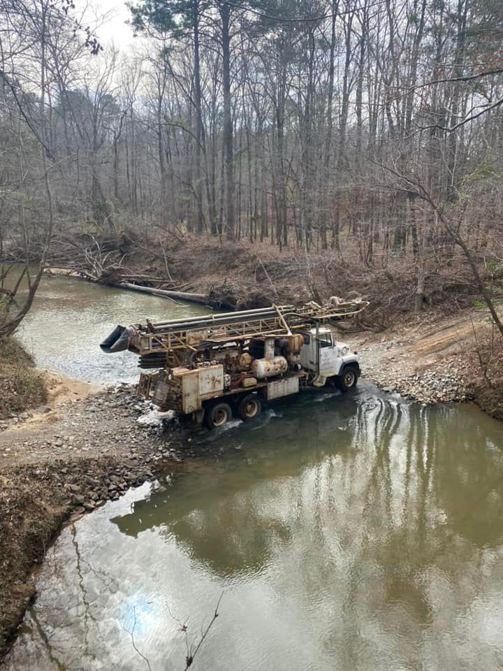 Truck with drilling equipment driving through a shallow, muddy creek; trees line the banks.