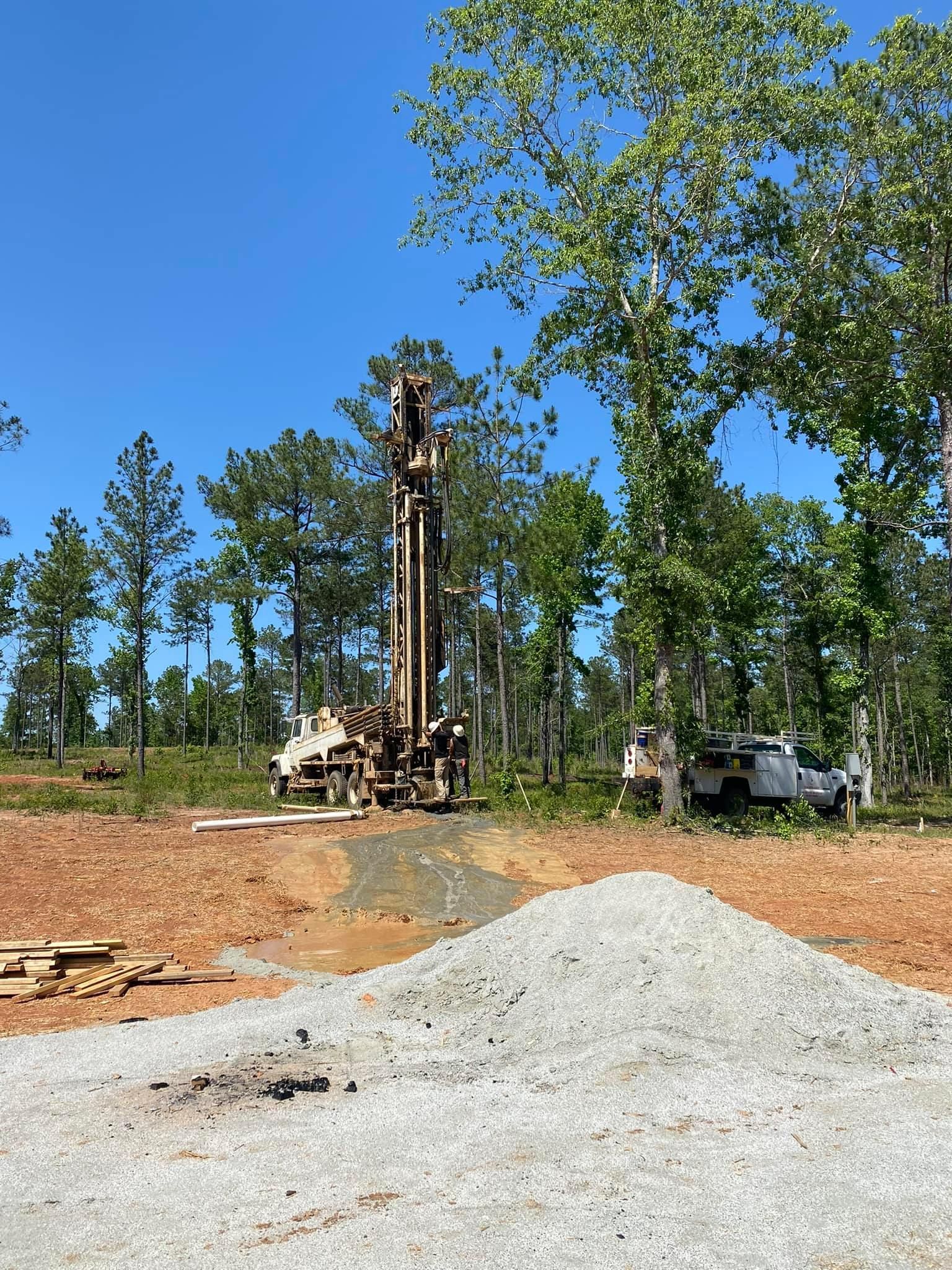 Drilling rig in a dirt clearing, trees in background, bright blue sky.
