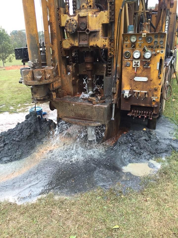 A yellow drilling rig in a grassy area is spewing water and dark mud.