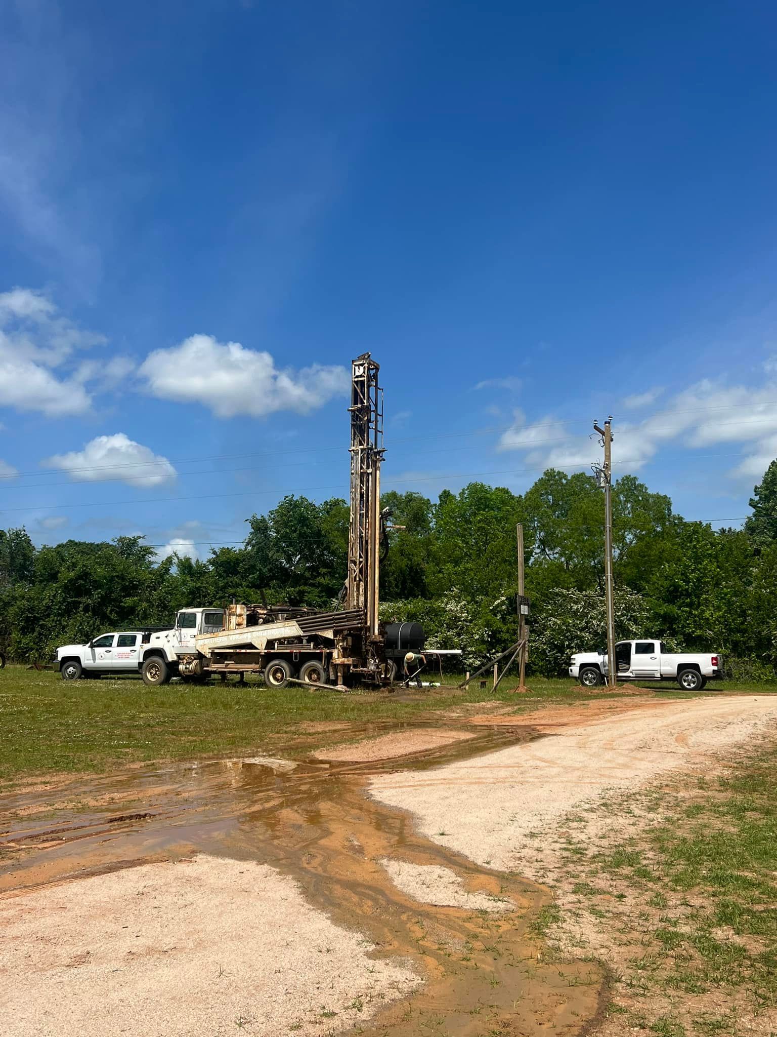 Drilling rig in a field, trucks, trees, and a bright blue sky.