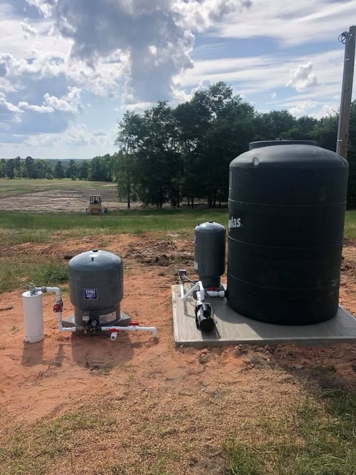 Water well system on a concrete pad: tank, filter, pressure tank, pipes, and pump in a field.