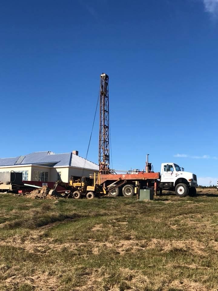 Drilling rig next to a house on a grassy field under a blue sky. White truck is parked nearby.