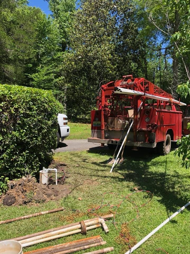 Red well-drilling truck next to a white truck on a grassy lawn with drilling equipment.