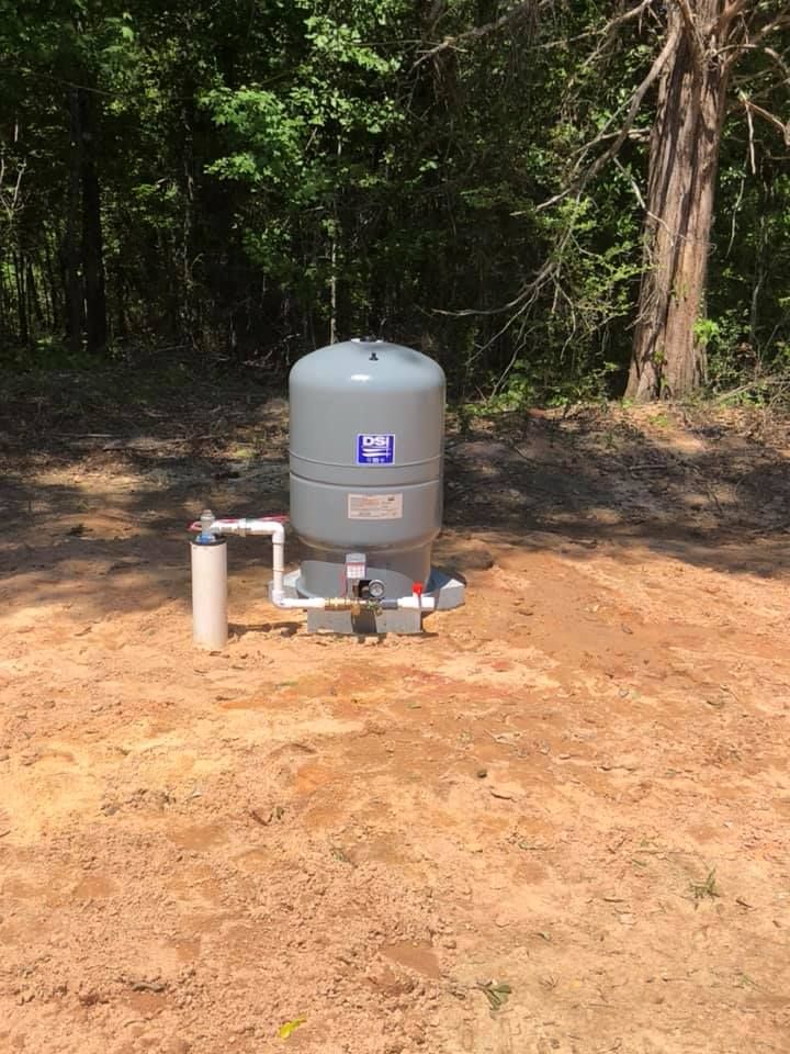 Gray water pressure tank and piping on dirt, surrounded by trees.