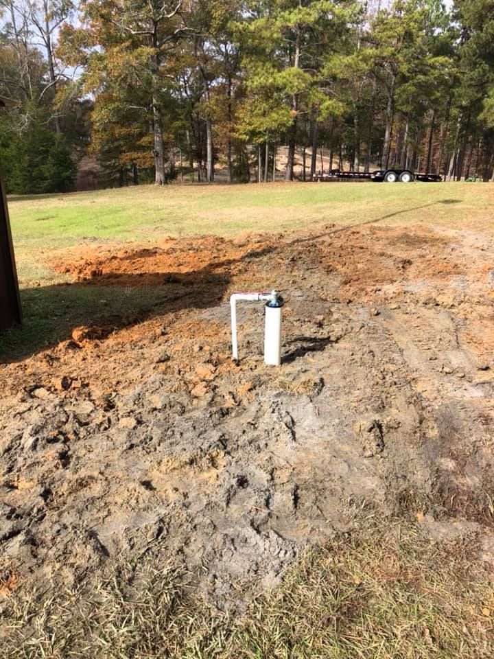 White well pipe in muddy soil, in a grassy field with trees in the background.
