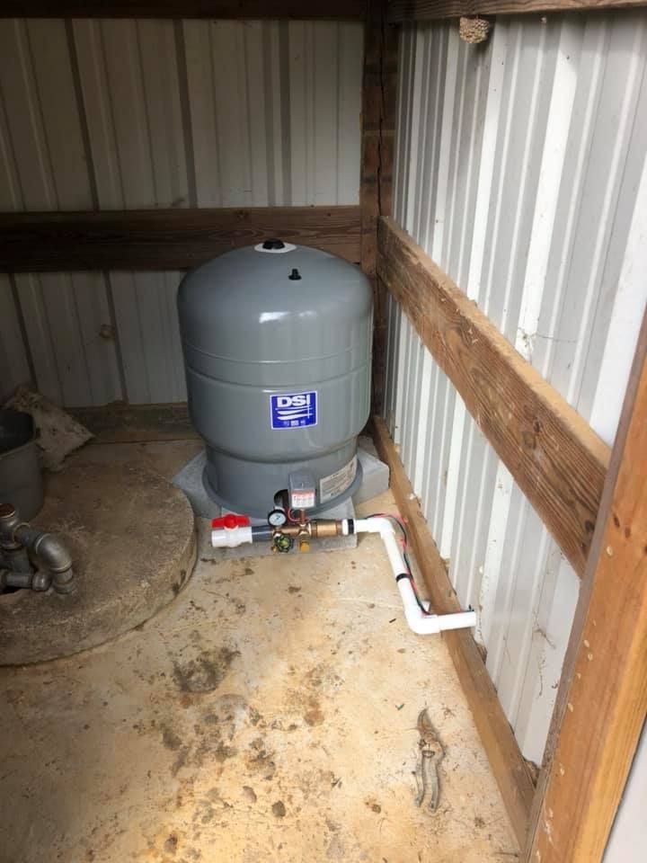 A water pressure tank inside a shed. The tank is gray, with pipes and a pressure gauge.