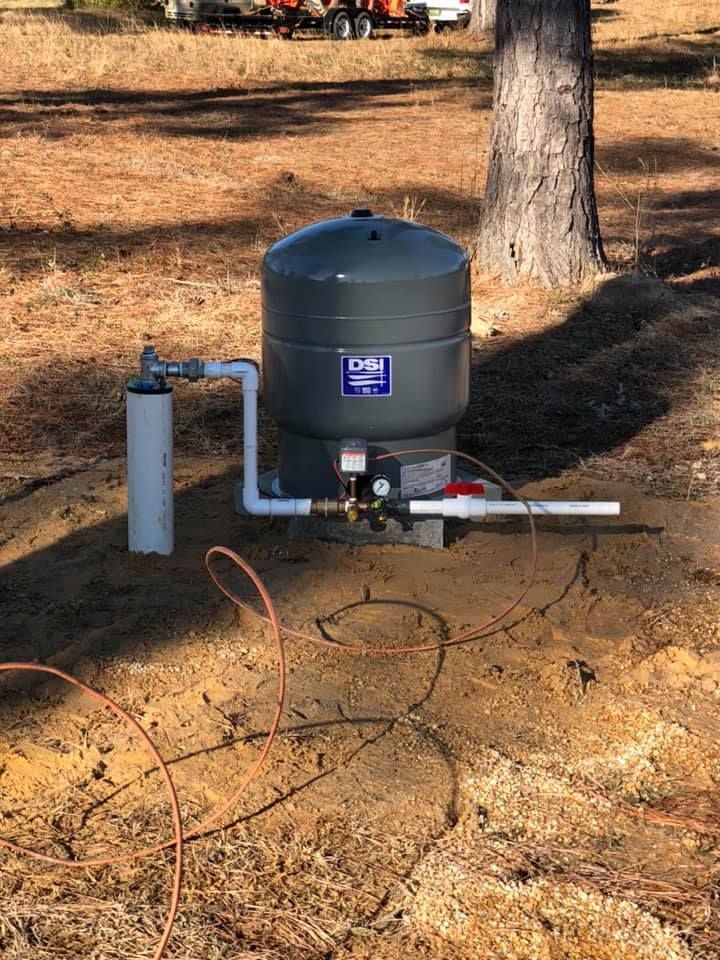 Well water pump system in a wooded area. Gray pressure tank and white pipes visible.