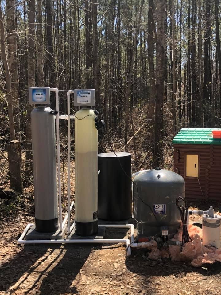Water filtration and well equipment outdoors in a wooded area, with storage tanks and pipes.