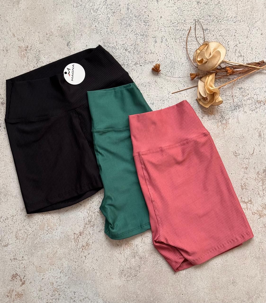 Tres pantalones cortos doblados, de color negro, verde y rosa, sobre una superficie clara con textura y flores secas.