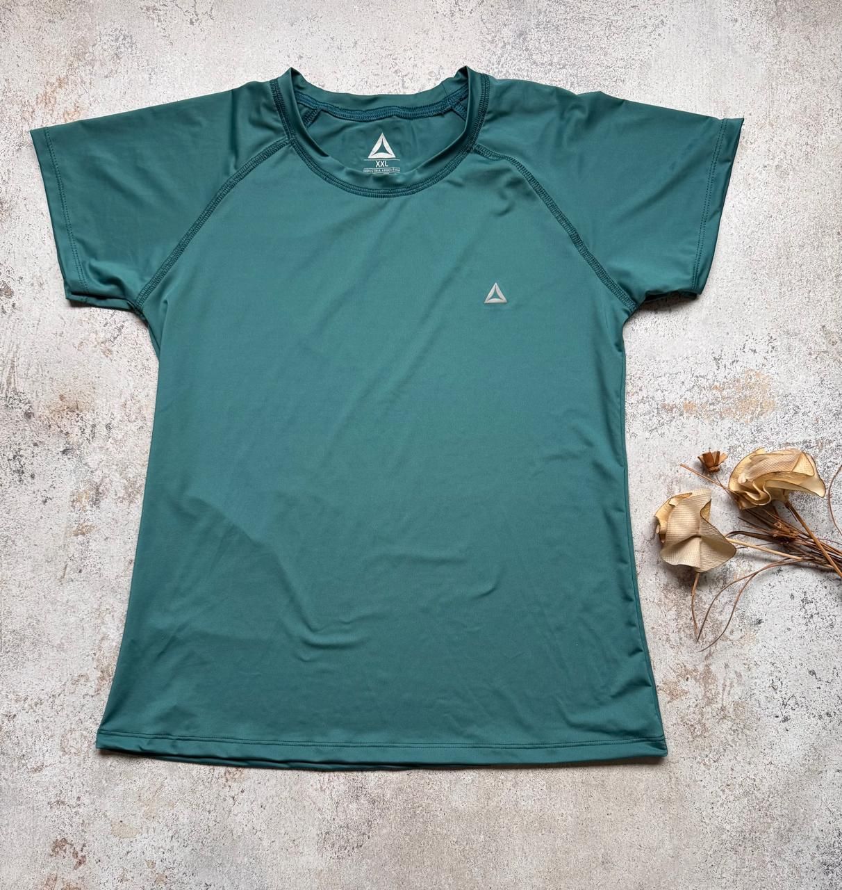 Camiseta deportiva de manga corta color verde azulado, extendida sobre una superficie clara con textura y flores secas cerca