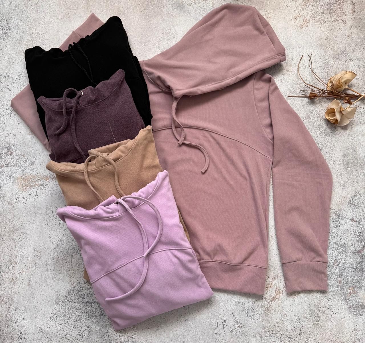 Cinco sudaderas con capucha dobladas, en negro, ciruela, tostado, lavanda y rosa empolvado, sobre un fondo claro con textura.