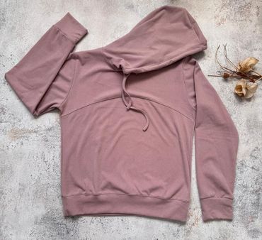 Sudadera con capucha color malva y manga a juego extendidas sobre una superficie gris texturizada con flores secas