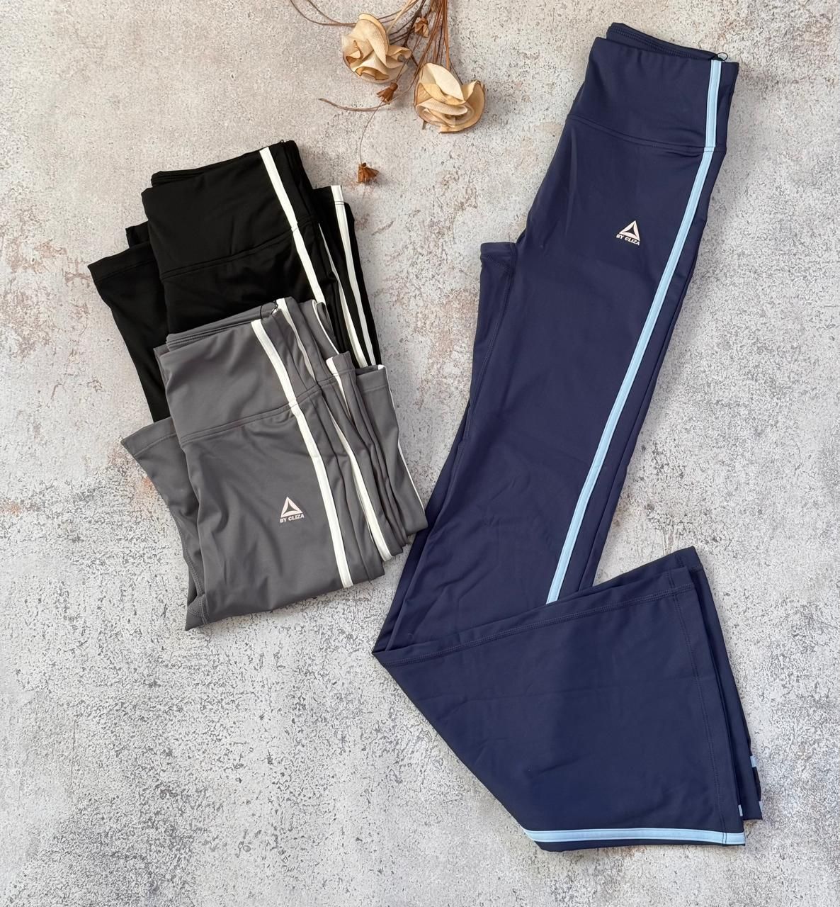 Pantalones deportivos negros, grises y azul marino, doblados y con rayas blancas, sobre una alfombra clara.
