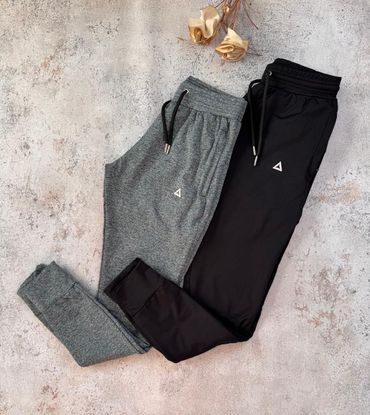 Pantalones deportivos grises y negros extendidos sobre una superficie de piedra clara, con un par de coleteros dorados cerca