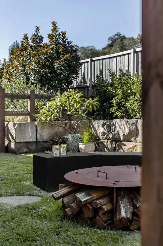 Backyard Fire Pit With Stacked Firewood, Grass, Stone Wall, and Trees in Sunlight — Billabong Pool Co in Smeaton Grange, NSW