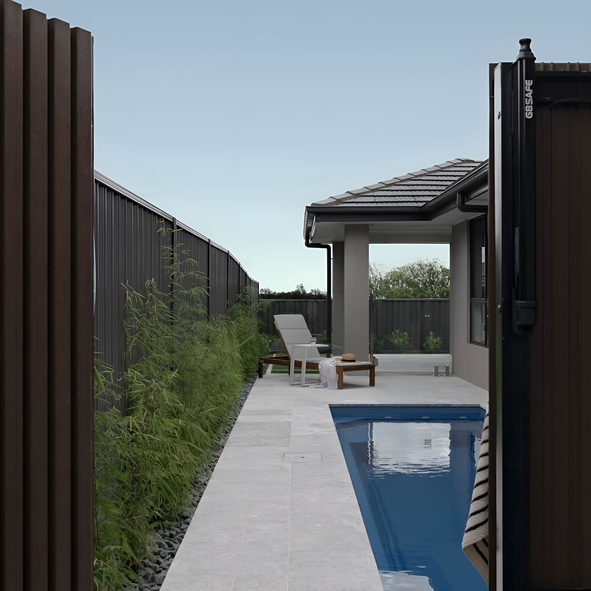 A narrow backyard with a pool and lounge chair. Dark brown fence and gate — Billabong Pool Co in Smeaton Grange, NSW