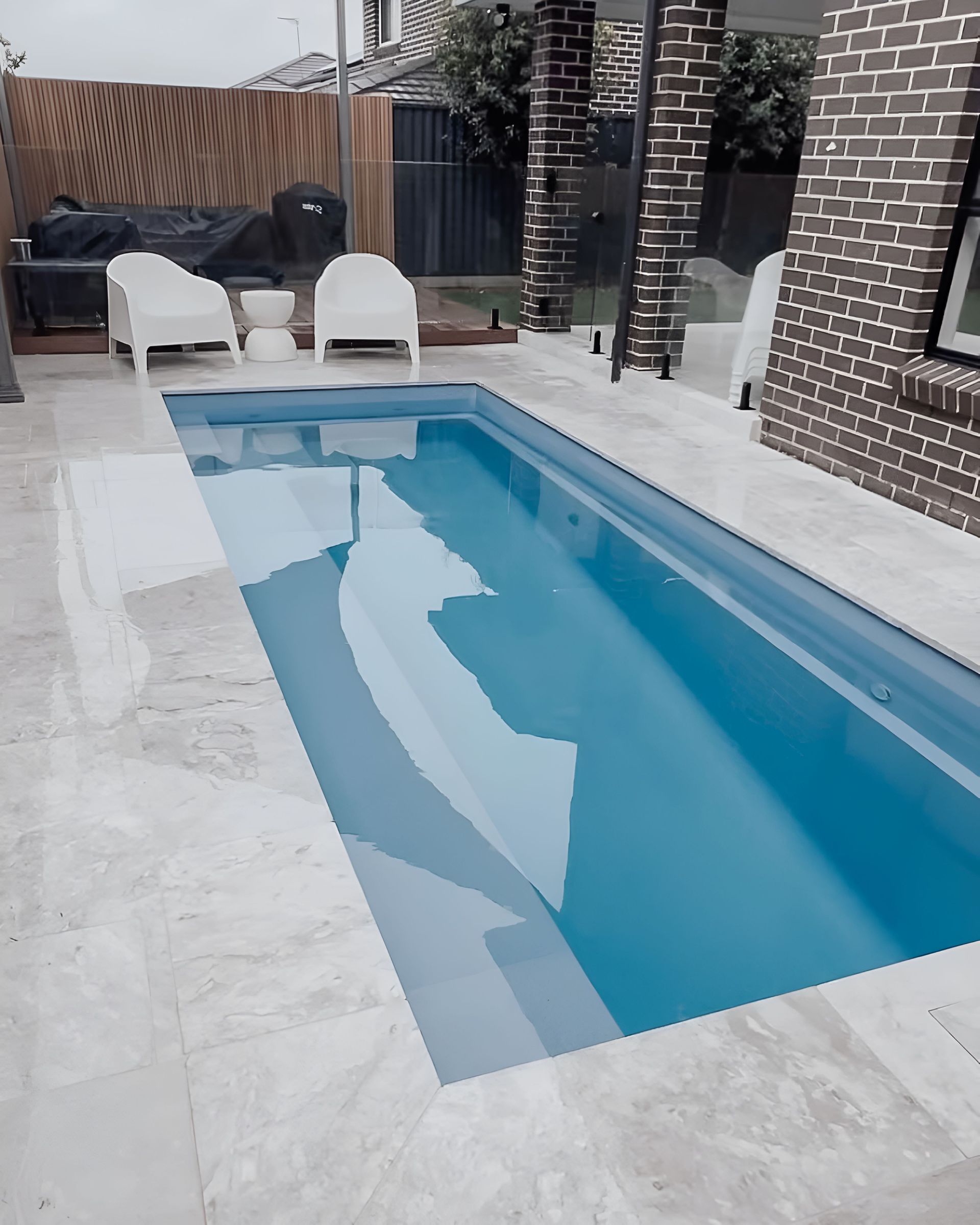 Narrow Backyard Swimming Pool Beside a Brick Patio With White Lounge Chairs and a Grill — Billabong Pool Co in Wollongong, NSW