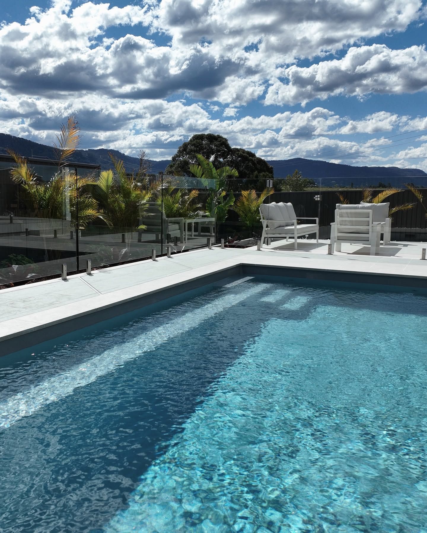 Backyard Pool With White Lounge Chairs, Mountains in the Distance, and a Cloudy Sky — Billabong Pool Co in Wollongong, NSW