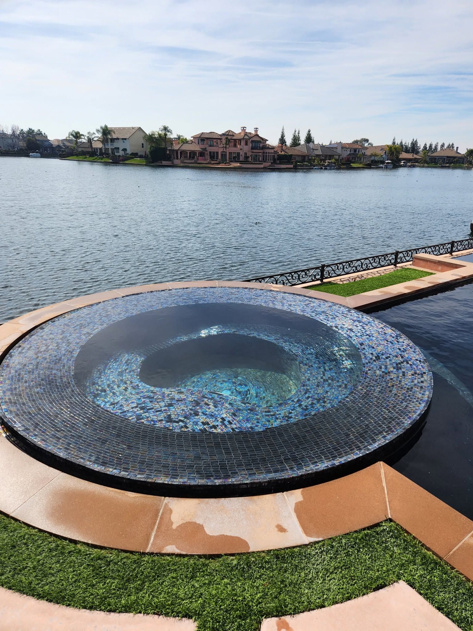 Curved mosaic fountain by a lake, with buildings and a blue sky in the distance.