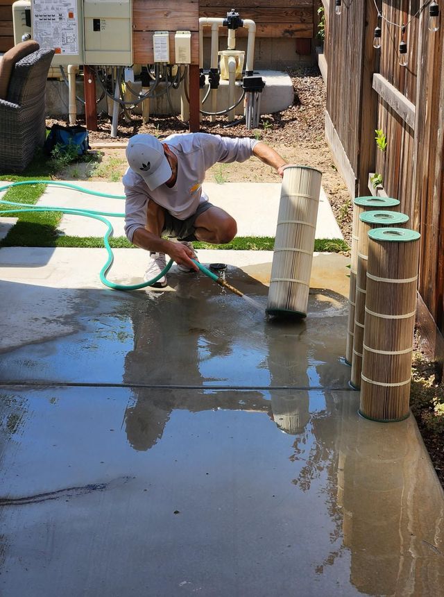 Worker crouching with a hose on wet concrete beside a wooden fence and a stone post.