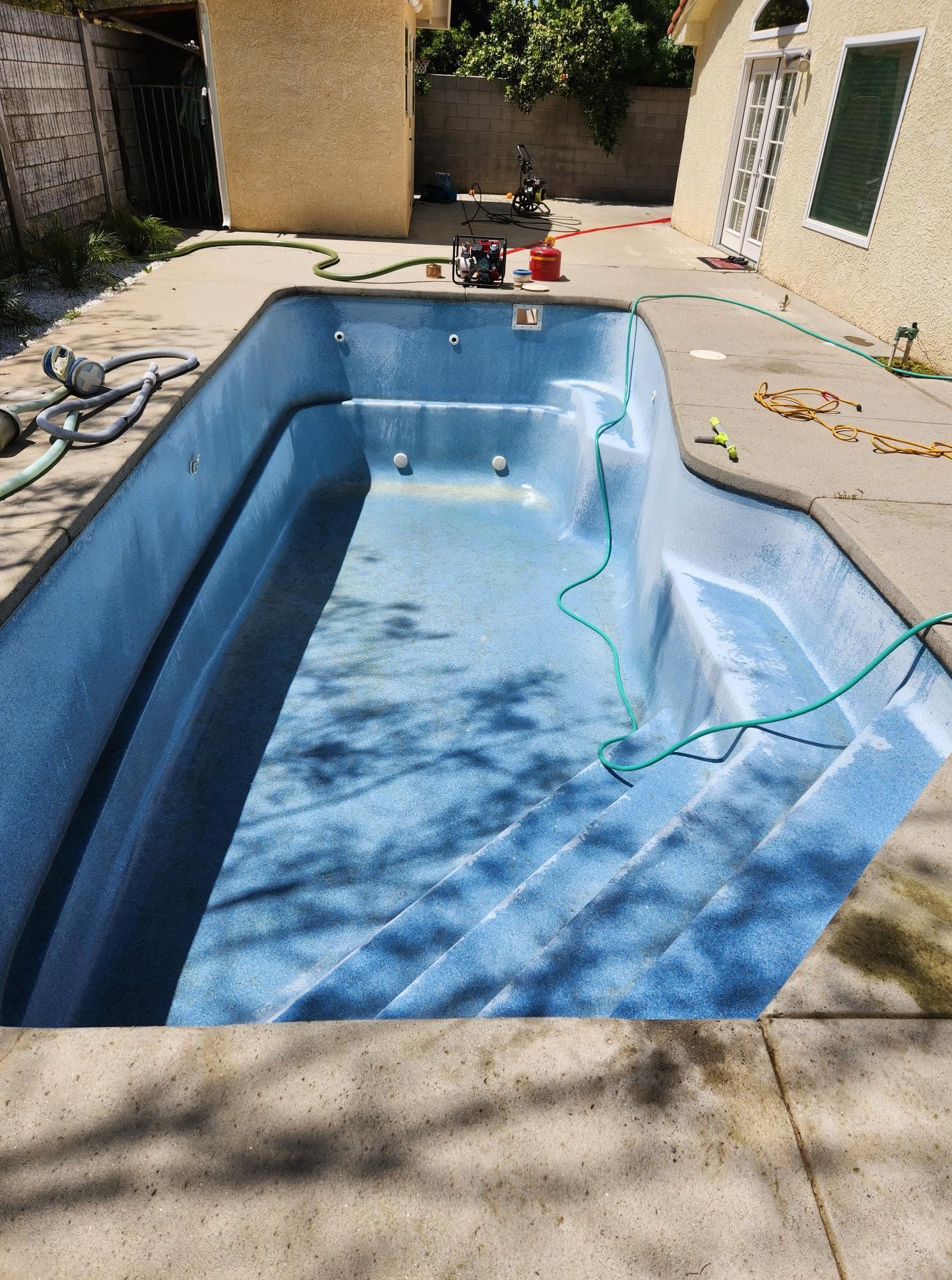 Empty blue swimming pool with a hose draped inside and cleaning tools on a sunny patio