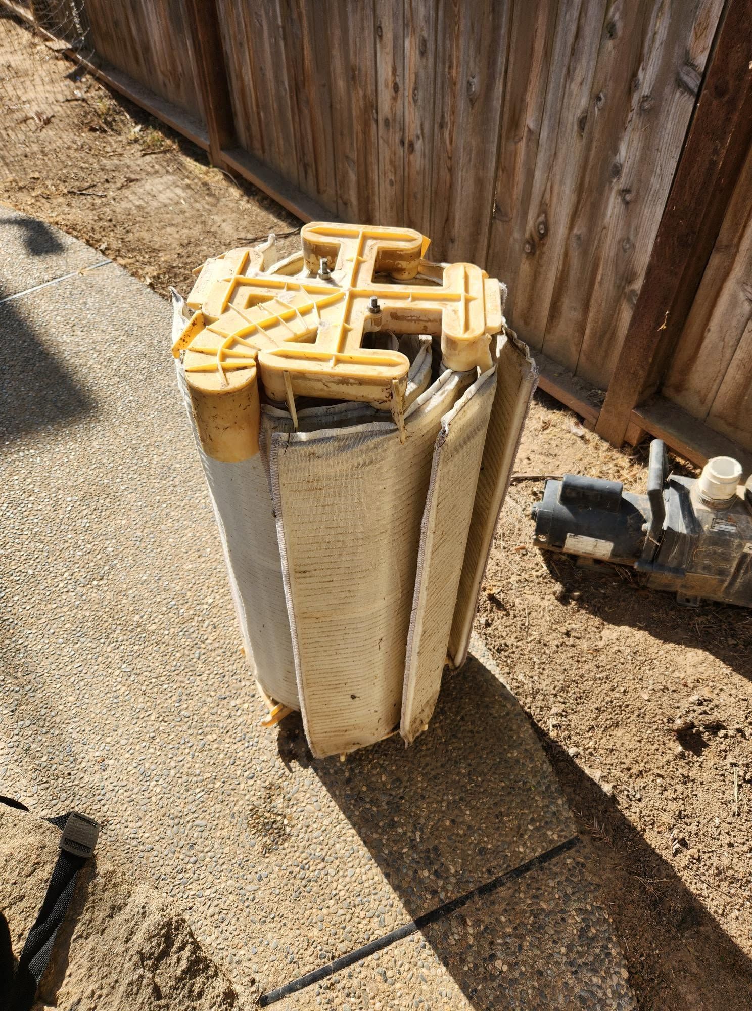Stacked beige foam and cardboard panels beside a wooden fence on gravel, with a small machine in the background.