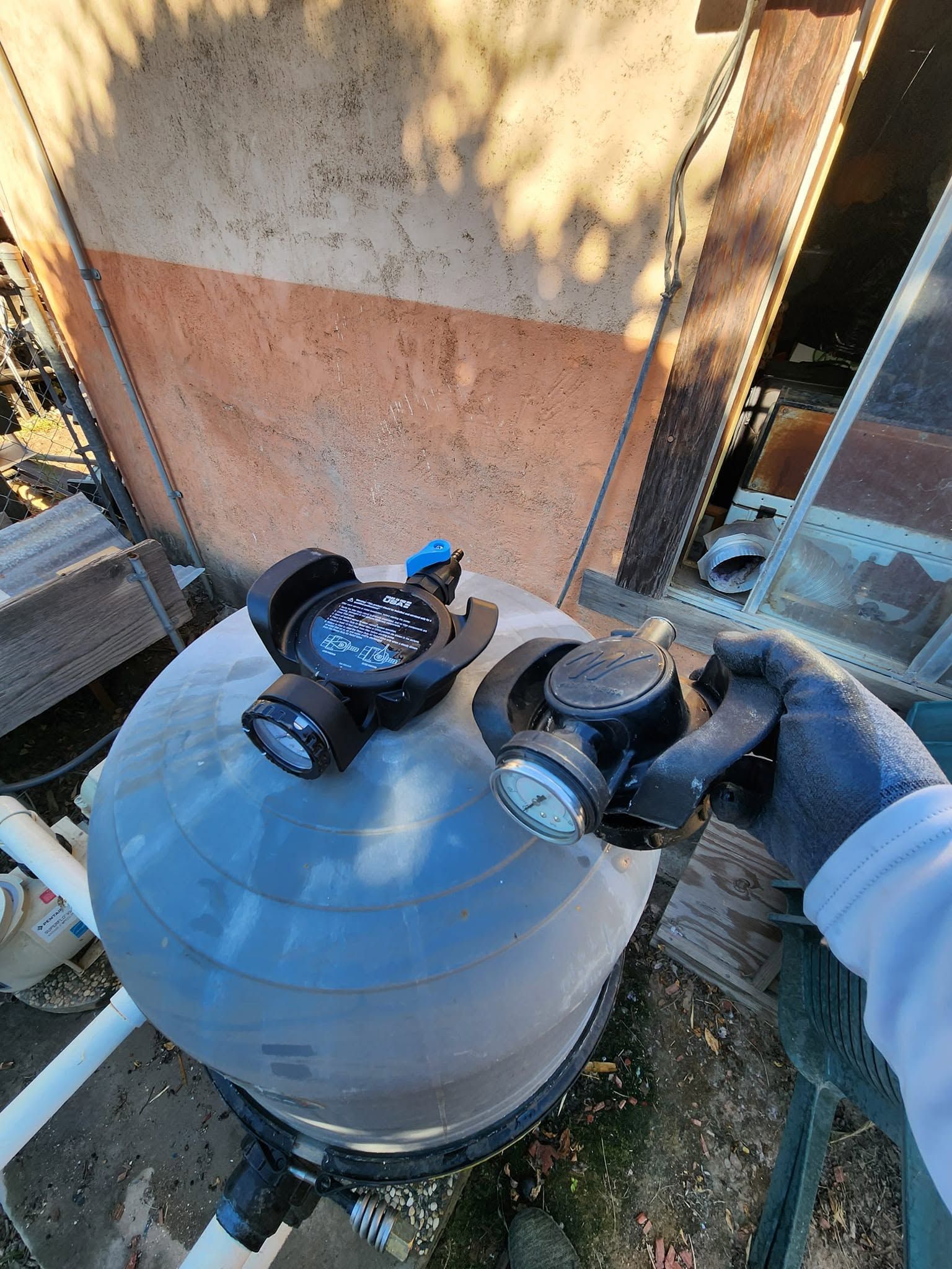 Gloved hand holding a large gray water filter tank with black fittings outdoors near a wall