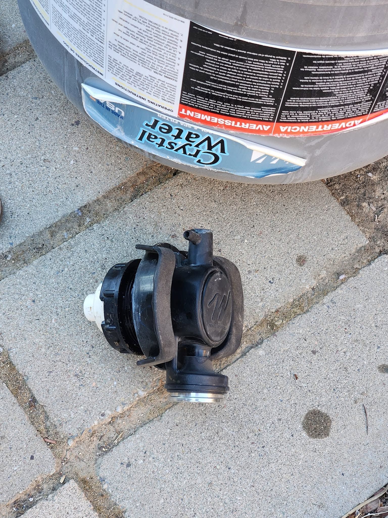 A metal and black valve-like part on a concrete floor beside a large container with warning labels.