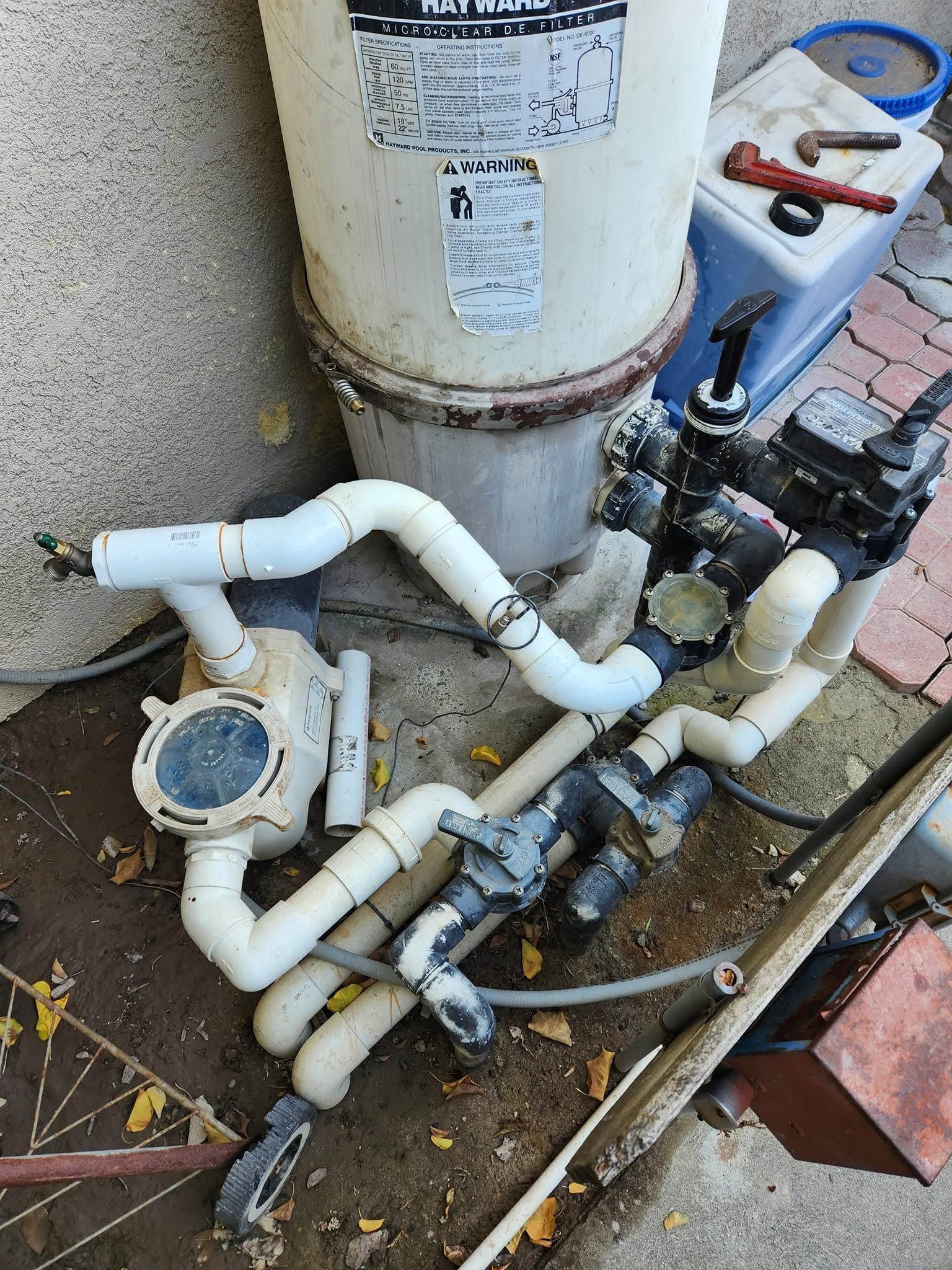 Outdoor pool plumbing with white pipes, valves, and pump equipment beside a beige tank and brick patio.
