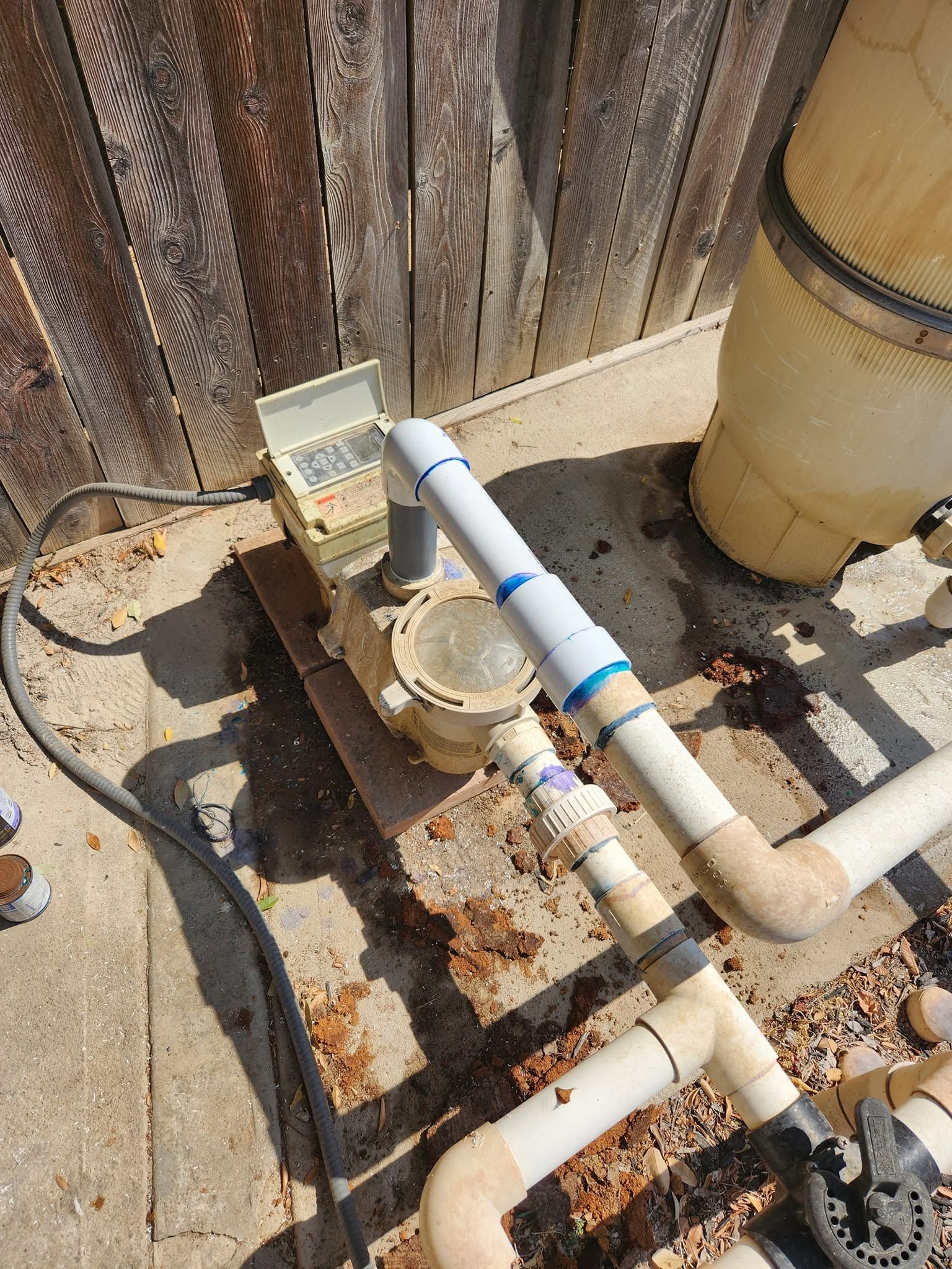 Outdoor pool equipment with white PVC pipes, pump, and rust stains on a concrete pad beside a fence