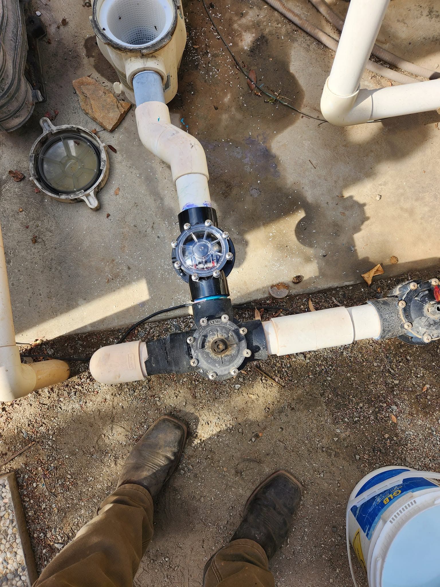 Underground PVC plumbing pipes and valves in a dirt utility area, with a person’s boots visible.