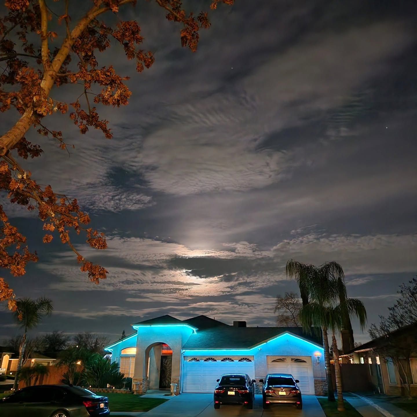 Moonlit suburban house with turquoise lights, two cars in the driveway, and cloudy night sky